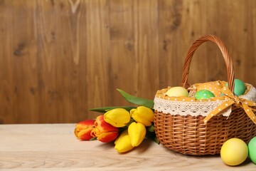 Easter eggs in wicker basket and beautiful tulips on wooden table, space for text