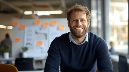 Fototapeta premium smiling man in blue sweater sits at desk in modern office, surrounded by creative materials and bright atmosphere