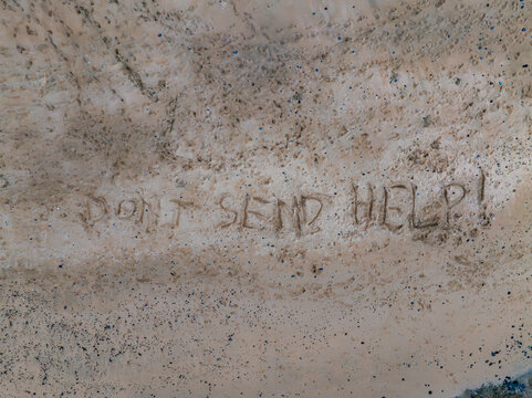 A sandy area features the message 'DON'T SEND HELP ' etched prominently, surrounded by scattered pebbles and debris, suggesting a coastal setting.