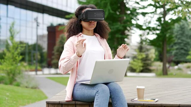 African american female student works using VR glasses in virtual reality simulator sitting on bench near university. Black woman uses gestures to control computer programs flips through virtual pages