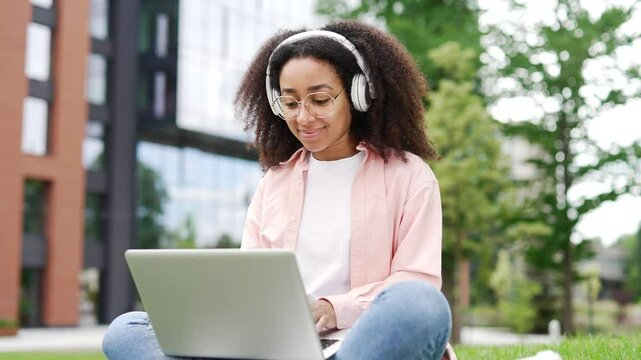 African american female student works on laptop sitting on lawn in campus space near university building. Young black woman in headphones learns remotely, listens to distance online seminar. Close up