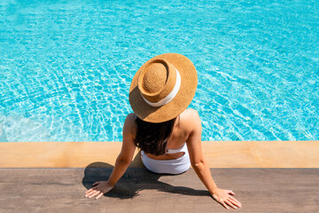 Happy traveler Asian female in white swimsuit and straw hat relaxing by the pool at hotel during her holiday. Summer and vacation, sunbathing wellness lifestyle concept. Copy space