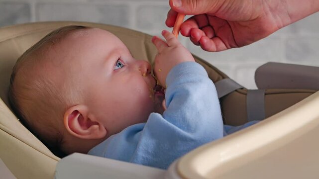 A small boy with a food-stained face eats porridge from a spoon. A woman's hand feeding a baby with a plastic baby spoon. Weaning a baby from the breast