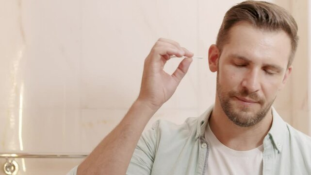 Smiling young man cleaning ear with cotton swab and looking to mirror at home bathroom
