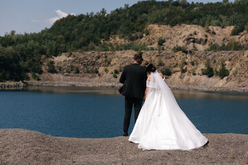 a couple standing with their backs to the camera on the shore of a pond. They look at the water, and behind them they can see a rocky shore and green trees on the hills.