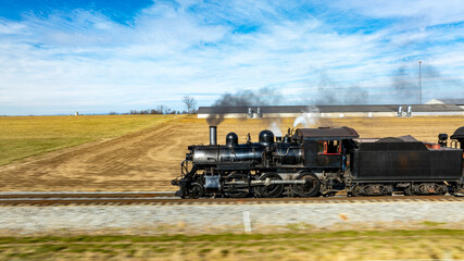 A vintage steam locomotive powers along the tracks, sending plumes of smoke into the blue sky. Surrounding fields and farmhouses enhance the picturesque countryside setting.