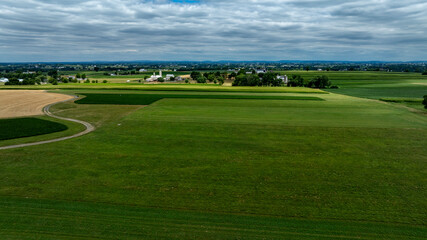 Aerial view captures extensive farmland with lush green fields and golden crops under a cloudy sky. Railroad tracks bisect the landscape, enhancing the serene rural setting.
