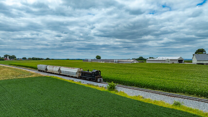 A vintage train moves steadily along tracks surrounded by lush green fields and barns. The overcast sky creates a tranquil atmosphere in the agricultural landscape.