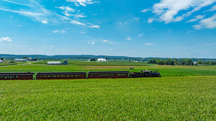A vintage train travels through expansive green fields under a bright blue sky. Farms and distant hills provide a picturesque backdrop, showcasing the beauty of rural life.