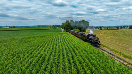 A steam locomotive speeds along railway tracks lined by vibrant green cornfields, under a partly cloudy sky. The countryside features rolling hills in the distance, creating a picturesque view.