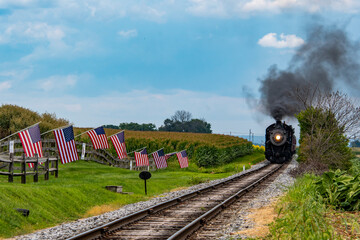 A steam locomotive puffs black smoke as it approaches a rural area. American flags decorate the landscape, adding a patriotic touch to the serene environment.