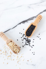 Black and white sesame seeds in wooden spoons on white marble table. Top view.
