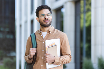 Student walking outside university campus with books in hand. Young man with backpack in casual...