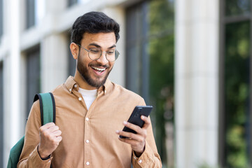 Man outside university campus with phone in hand celebrating successful exam results, student with...