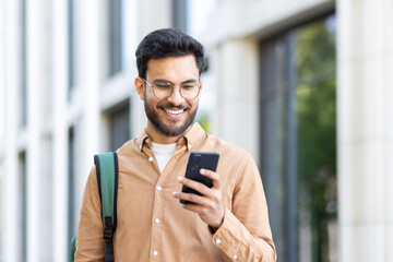 Young student with backpack on shoulder using app on smartphone. Man walking outside university...