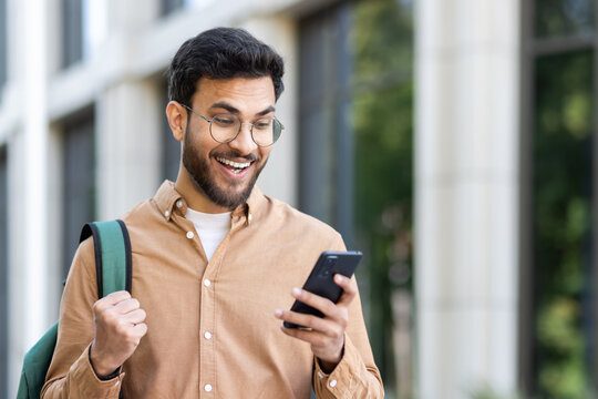 Man outside university campus with phone in hand celebrating successful exam results, student with backpack holding hand up in triumph gesture, received online notification on smartphone.