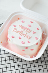 Birthday little cake with white cream cheese frosting decorated with happy birthday text and pink cream hearts on top. Trendy Korean bento cake in the white gift box on the white marble background
