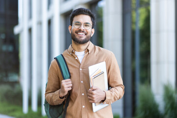 Portrait of a happy successful student with books and backpack outside the university campus. The man is smiling and looking at the camera, dressed in casual clothes.