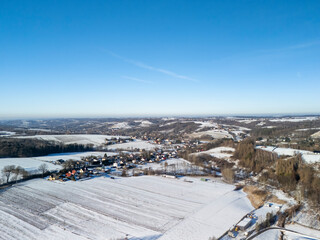 Fototapeta premium Aerial view of a snow-covered village surrounded by hills under a clear blue sky, showcasing winter serenity and natural beauty.