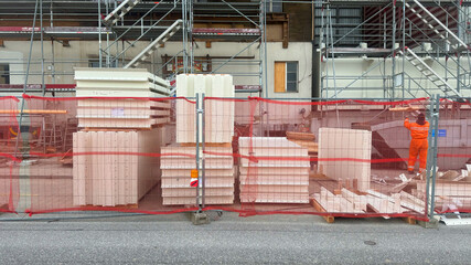 A construction site enclosed by red safety netting, with stacks of building materials neatly arranged in front. A worker in an orange protective suit is seen handling materials, while metal