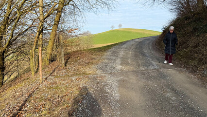 Obraz premium An elderly woman with a walking stick strolls along a winding gravel road in a peaceful countryside setting. The road curves gently around rolling green hills, creating a serene and scenic atmosphere.