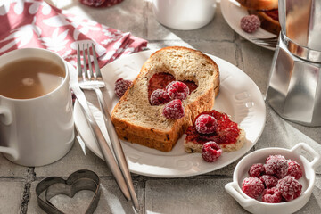 Toasted bread with heart-shaped cut and raspberry jam on tile background in bright morning light. Festive breakfast for Valentines day