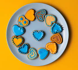 Valentine’s day composition with cookies in shape of hearts decorated by yellow and blue icing and sprinkles on blue plate against yellow background