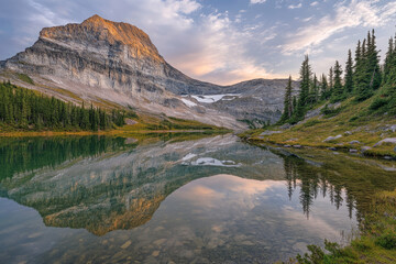 Fototapeta premium A tranquil mountain lake at sunrise with calm, glass-like reflections and soft pastel sky hues.