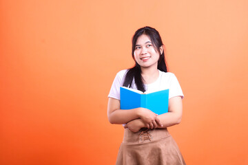 A woman with a blue book, looking up and to the right, dressed in white tee and brown skirt,isolated on orange background