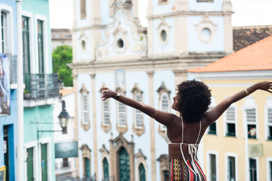Young black woman with afro hairstyle, back to the camera, arms raised. Against old houses in the background. Happy and empowered person.