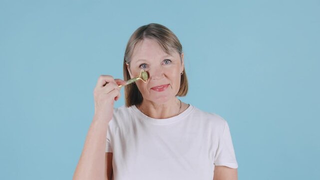 Woman using a jade roller for facial skincare against a light blue background while maintaining a cheerful expression and promoting self-care routines