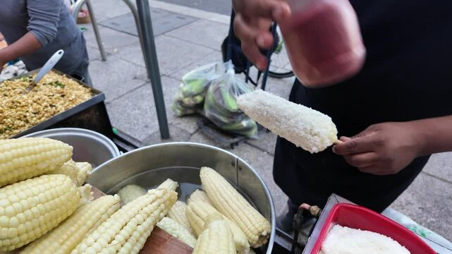 A man is holding a corn on a stick and is about to eat it. The corn is in a pot and there are other corn pieces in the pot