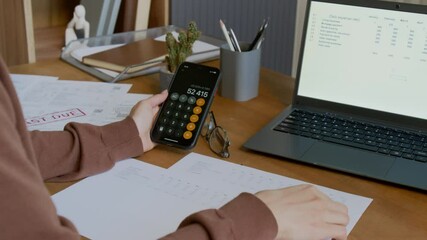 Medium shot of unrecognizable young female homeowner calculating domestic expenses on smartphone app, then circling numbers on invoice, while sitting at desk in front of laptop with budget planner