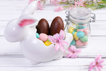 Easter decor on the white wooden table - white and pink bunny shaped vase with colorful and chocolate eggs, hyacinth pink flowers, jar with assorted eggs and a blooming branch on the background.