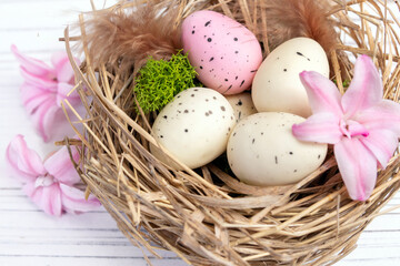 Easter decor on the white wooden table - nest with speckled pink and beige eggs, moss and feathers and sprinkled pink hyacinth flowers around.