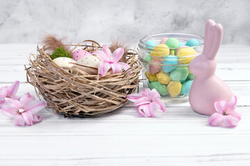 Easter decor on the white wooden table - nest with speckled pink and beige eggs, moss and feathers; glass jar with colorful chocolate egg candies and a figurine of ceramic pink bunny, sprinkled hyacin