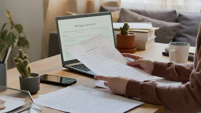 Medium closeup of anonymous young woman sitting at desk at home, reading through mortgage agreement on paper copy and file on laptop screen, while preparing to take out bank loan to buy house