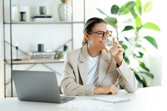 Positive caucasian businesswoman enjoying a refreshing break while drinking a glass of water, sitting at her desk in a modern office with a laptop, emphasizing wellness and work-life balance