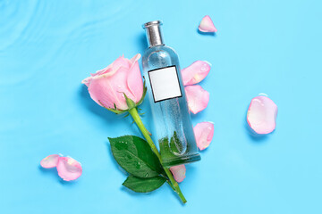 Bottle of perfume with beautiful pink rose and petals in water on blue background