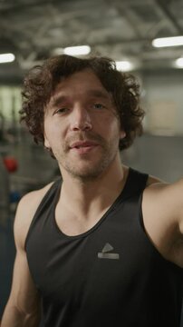 Athletic man looking into camera, waving and talking as recording social media vlog or video calling in gym. Vertical UGC clip