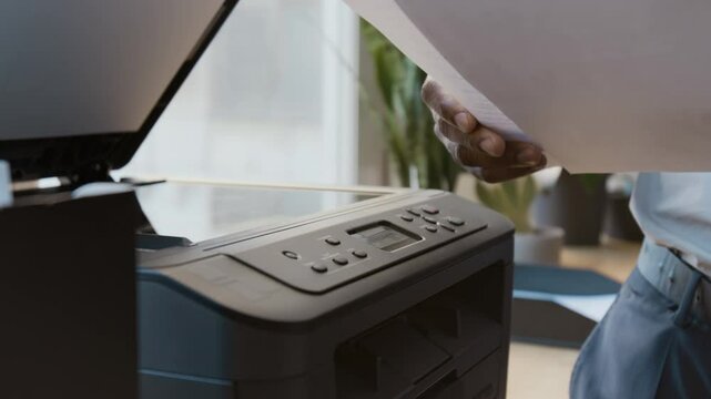 Medium closeup of midsection of black male office worker walking up to photocopier, opening cover, putting down document and pressing start button for copying or scanning