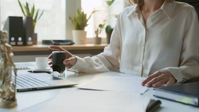 Medium midsection shot of unrecognizable female paralegal or legal assistant in white blouse stamping document copies with official seal, while managing paperwork at law firm or notary office