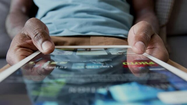 Closeup of hands of unrecognizable African American man sitting on couch, browsing through TV shows on streaming service application on digital tablet, while choosing movie to watch