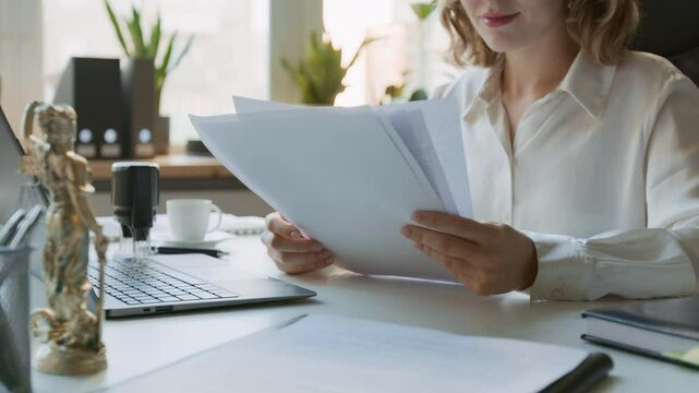 Medium midsection shot of smiling anonymous young female attorney, wearing white blouse, sitting at law firm or notary office, studying paper copies of legal documents. Lady Justice statuette on desk
