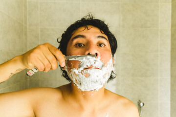 Photograph of young latin man shaving with razor and shaving cream in front of the mirror. Concept...