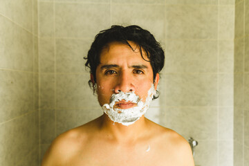 Photograph of young latin man shaving with razor and shaving cream in front of the mirror. Concept...