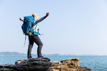 Obraz premium A senior woman stands triumphantly on a rocky shoreline, arms outstretched toward the ocean. Wearing a backpack and hat, she embraces the freedom, adventure, and beauty of the coastal landscape.
