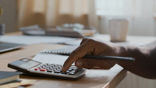 Closeup of hands of unrecognizable black man adding up living costs and utility charges on calculator, then writing down sums in notebook, while managing family budget