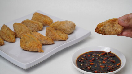 One hand is holding fried tahu walik aci, there is a plate of tofu walik with soy sauce and chili sauce beside it, placed on a white base and background (isolated white).
