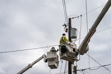 Line crew workman in hydraulic lift bucket high up working on power lines at wood utility pole,...